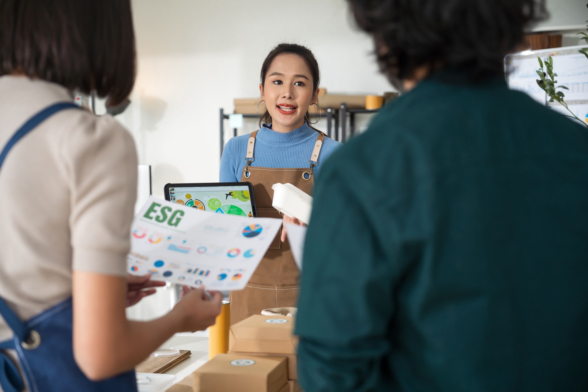 Asian business meeting features a female entrepreneur in apron showcasing eco-friendly, reusable product. The discussion centers on environmental protection and building a sustainable business model